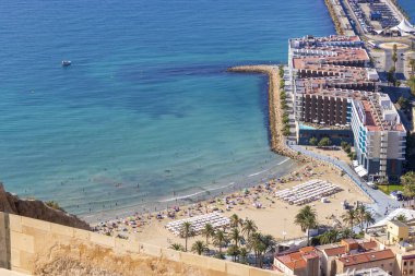 Playa del Postiguet 'in Alicante' deki hava manzarası açık mavi gökyüzünün altında güneşlenen kumlu plajları ve palmiye ağaçlarını gösteriyor..