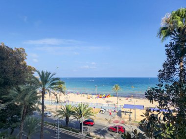 Alicante Spain - 12 Temmuz 2025: Playa del Postiguet plajının renkli palmiye ağaçları ve Akdeniz kıyısında güneşli havanın keyfini çıkaran insanların manzarası.