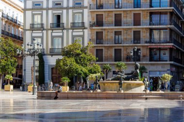 Valencia İspanya - 13 Temmuz 2025: Plaza de la Virgen 'deki Fuente del Turia' da güneşli bir gün canlı ağaçlar ve tarihi binalarla çevrili çarpıcı bir çeşme sergileniyor.