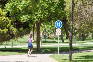 Valencia İspanya - 13 Temmuz 2025: Bir kadın ağaçlarla çevrili canlı yeşil bir parkta koşuyor ve yaya geçitlerini destekleyen tabelalar gösteriyor.