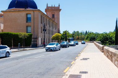 Valencia İspanya - 13 Temmuz 2025: Şehrin canlı atmosferini gözler önüne seren tarihi binalar ve yeşillik yakınlarındaki bir caddede arabaları tasvir eden şehir manzarası.