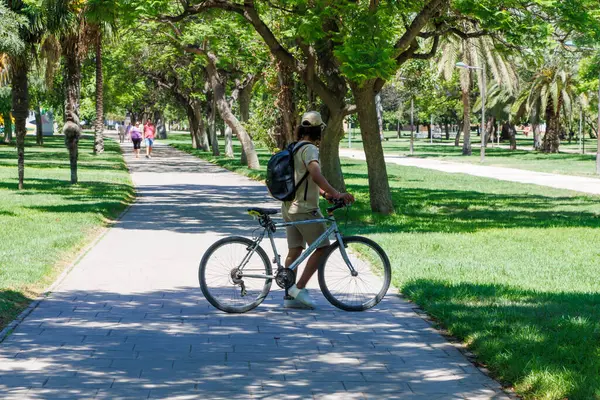 Valencia İspanya - 13 Temmuz 2025: Ağaçlarla çevrili yemyeşil bir parkta bisikletli genç adam güneşli günün tadını çıkarıyor.