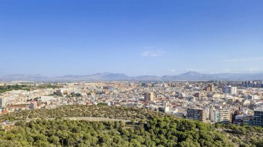 Açık mavi gökyüzünün altında yeşil tepelere kurulmuş canlı binalarıyla Alicante şehrinin panoramik manzarası kentsel yaşamı ve doğal güzellikleri gözler önüne seriyor..