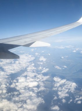 Açık mavi gökyüzüne ve kabarık bulutlara karşı bir kanat bulunan bir uçak penceresinden görüntü. Gündüz yolculuk ve havacılık temaları için mükemmel.