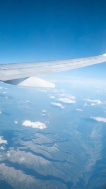 Ön planda görünen uçak kanadı, yüksek irtifada güneşli bir günde aşağıdaki dağları ve bulutları nefes kesici bir şekilde gösteriyor..