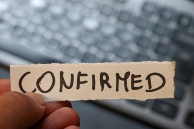 Bir el, üzerinde CONFIRMED sözcüğünün yazılı olduğu bir kağıt parçasını bir bilgisayar klavyesinin bulanık arkaplanının doğruluğu ve tamamlanmasını simgeleyen arkaplanına karşı tutar.