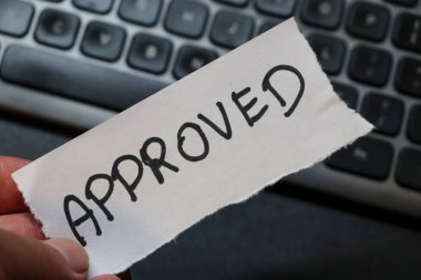 Ofis ortamında görevlerin veya projelerin başarıyla tamamlandığını sembolize eden bir el bilgisayarda bir klavyenin üzerinde bir APPROVED notu tutuyor. İşle ilgili bağlamlar için ideal.