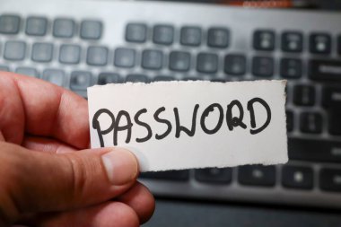 Modern teknolojide güvenlik ve dijital gizliliği vurgulayan bir bilgisayar klavyesinin arka planına karşı PASSWORD sözcüğüyle bir notu tutan bir el..