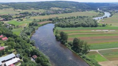 Mavi gökyüzünün altında geniş bir nehrin tarlalar ve otlaklar arasında aktığını gösteren drone görüntüsü.
