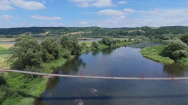 Yeşil, yemyeşil bir vadide geniş bir nehri geçen kırmızı asma köprünün hava görüntüleri..