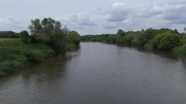 Bereketli yeşil bankalar ve bulutlu bir gökyüzünün altındaki uzak tarım arazileri boyunca kıvrılan sakin bir nehrin hava videosu..