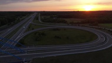 Günbatımında modern otoban yonca yaprağı kavşağının insansız hava aracı görüntüsü, turuncu gökyüzü aydınlatma yol ağı ve kırsal alan.