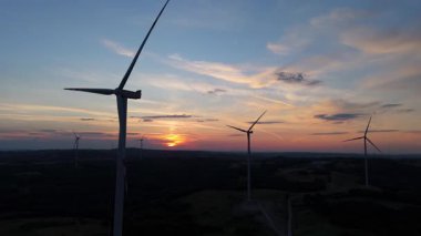 Kırsal tepelerdeki rüzgar türbinlerinin hava günbatımı görüntüsü, renkli akşam gökyüzü ile yenilenebilir enerji çiftliği.