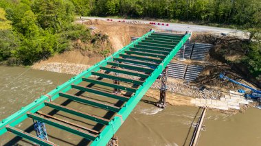 Aerial view of a partially constructed green steel bridge over a river surrounded by lush greenery. Heavy machinery is visible nearby indicating ongoing construction and development.
