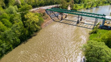 Yeşilliklerle çevrili bir nehir üzerinde kısmen inşa edilmiş bir köprünün havadan görünüşü. İnşaat makineleri devam eden altyapı gelişimini gösteren görünür bir özelliktir.