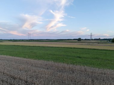 Alacakaranlıkta geniş yeşil alanlar gösteren güzel bir manzara. Elektrik direkleri rengarenk gökyüzüne karşı dimdik durur kırsal kesimin sükunetini ve cazibesini gösterir..