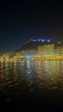 Alicante 'nin rıhtım boyunca aydınlatılmış binaları sergilediği göz kamaştırıcı gece manzarası. Manzaralı arka planda sakin sularda yansıyan bir dağ ışığı var..