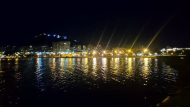 Night view of Alicante waterfront illuminated by city lights and reflections on water. The scene captures urban life by the sea showcasing the beauty of this coastal location.