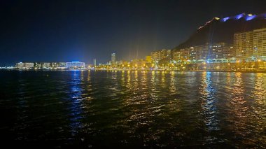 Stunning nighttime view of Alicante beach featuring illuminated waterfront buildings reflecting on water under a starry sky. A serene atmosphere perfect for travel promotions.