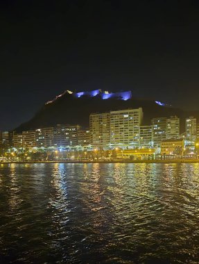 Sahile bakan aydınlık Alicante şatosunun gece manzarasında kıyı boyunca modern binalarla çevrili sakin denizde renkli yansımalar var..