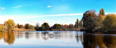 Durgun suların üzerinde nefes kesici bir manzara beliriyor, ağaçlar kenarları çerçeveliyor ve sonbahar renkleri akşam ışığında açık mavi gökyüzüne karşı güzel bir kontrast oluşturuyor..