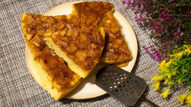 Tatlı bir karamelize pastanın dilimleri bir tabakta ustalıkla düzenlenir, arka plandaki renkli çiçeklerle tamamlanmış zengin altın renkleri ve davetkar dokuları sergilenir..