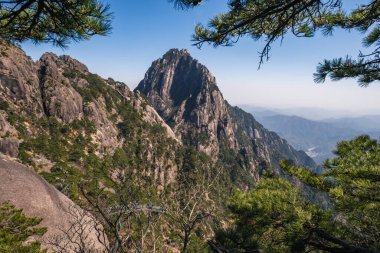 Çin 'deki Huangshan Dağları' nın nefes kesici manzarası. Engebeli zirveler, bereketli çam ormanları ve uzaktaki sisli bir vadiyle çerçevelenmiş gökyüzünü deler. Bu görüntü doğanın saf güzelliğini ve ihtişamını yakalar, derin düşüncelere ve meraka davetiye çıkarır..