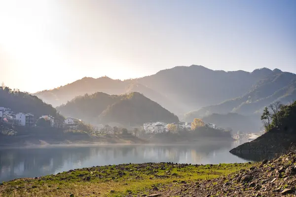 Sakin bir göle yansıyan sakin bir dağ manzarası. Güneşin doğuşu dağları ve köyü altın bir ışıkla kaplıyor ve güzel bir manzara yaratıyor. Sakin su, çevredeki yeşillikleri ve yumuşak tepeleri yansıtıyor. Huzurlu ve huzurlu bir ortam yaratıyor. 
