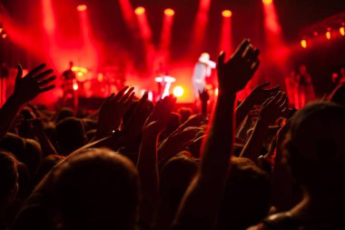 Festivaldeki konser kalabalığı, birçok insanın elleri sahnede müzik çalan müzisyenleri alkışlıyor, kırmızı arka ışık