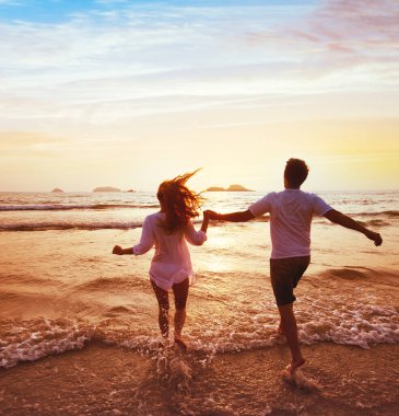 Balayı seyahatindeki mutlu çift, romantik rüya kumsal tatili, mutluluk geçmişi, gün batımında denize koşan kadın ve erkeğin siluetleri.