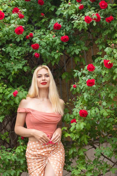 長いブロンドの髪と庭でバラの花の近くにピンクの服を着て赤い唇を持つ美しい女性のポーズ ライトピンクのトップとスカートを着用 — ストック写真