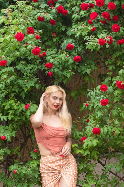 美丽的女人 金发碧眼 身穿粉色衣服 在花园里摆出绽放的玫瑰 穿浅粉上衣和裙子 — 图库照片