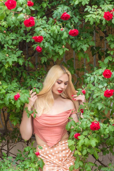 長いブロンドの髪と庭でバラの花の近くにピンクの服を着て赤い唇を持つ美しい女性のポーズ ライトピンクのトップとスカートを着用 — ストック写真