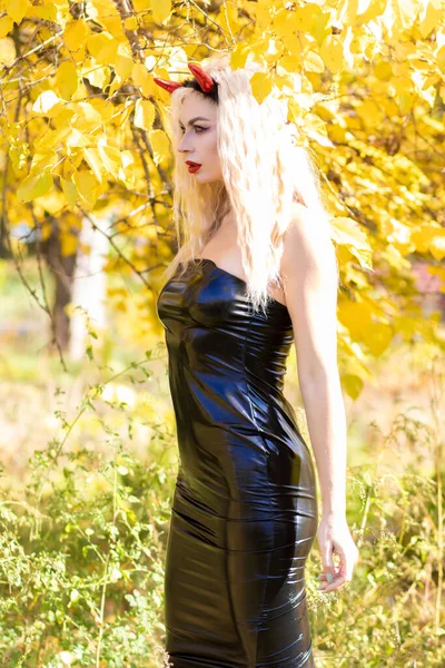 美しい金髪の女性は 自然の中で悪魔の衣装を着ています ブラックラテックスタイトなドレスとホーン ソフトフォーカス ハロウィーンコンセプト — ストック写真