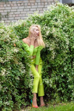 Sarı-yeşil takım elbiseli, uzun saçlı, güzel bir sarışının uzun boylu portresi ve çiçekli yeşil çalıların arka planında parlak yüksek topuklu ayakkabılar.