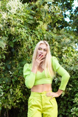 Sarı-yeşil takım elbiseli, uzun saçlı güzel bir sarışının portresi çiçekli yeşil çalıların arka planında.