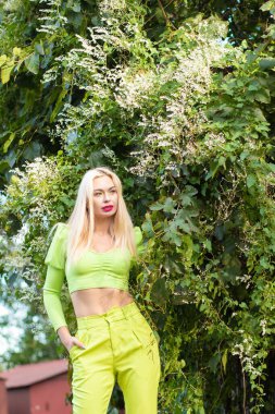 Sarı-yeşil takım elbiseli, uzun saçlı güzel bir sarışının portresi çiçekli yeşil çalıların arka planında.