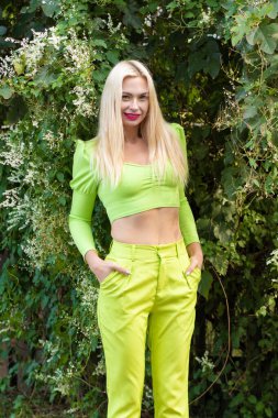 Sarı-yeşil takım elbiseli, uzun saçlı güzel bir sarışının portresi çiçekli yeşil çalıların arka planında.