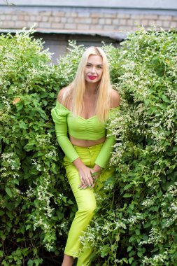 Sarı-yeşil takım elbiseli, uzun saçlı, güzel bir sarışının uzun boylu portresi ve çiçekli yeşil çalıların arka planında parlak yüksek topuklu ayakkabılar.