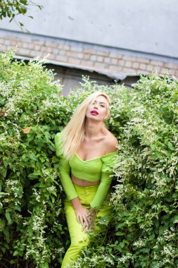 Sarı-yeşil takım elbiseli, uzun saçlı, güzel bir sarışının uzun boylu portresi ve çiçekli yeşil çalıların arka planında parlak yüksek topuklu ayakkabılar.