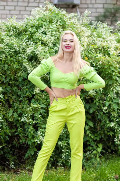 Sarı-yeşil takım elbiseli, uzun saçlı, güzel bir sarışının uzun boylu portresi ve çiçekli yeşil çalıların arka planında parlak yüksek topuklu ayakkabılar.