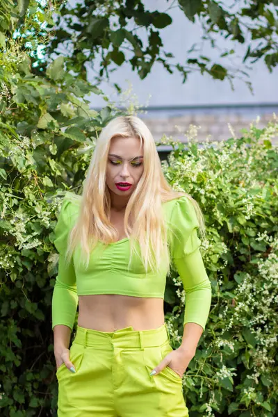 Sarı-yeşil takım elbiseli, uzun saçlı güzel bir sarışının portresi çiçekli yeşil çalıların arka planında.