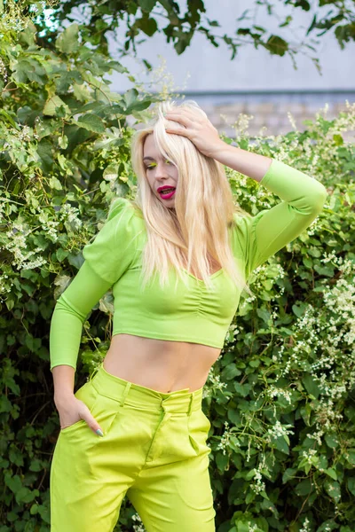 Sarı-yeşil takım elbiseli, uzun saçlı güzel bir sarışının portresi çiçekli yeşil çalıların arka planında.