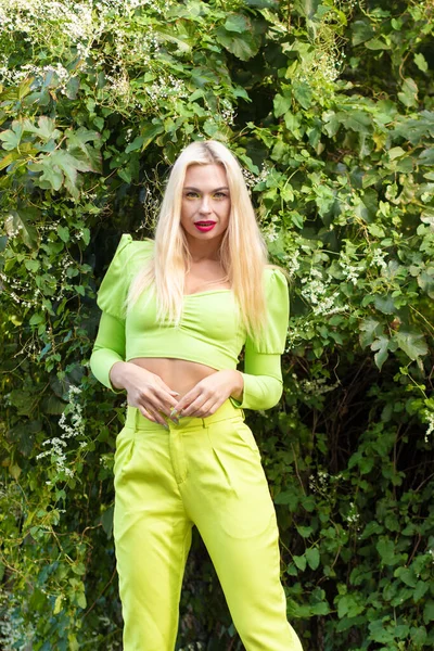 Sarı-yeşil takım elbiseli, uzun saçlı güzel bir sarışının portresi çiçekli yeşil çalıların arka planında.