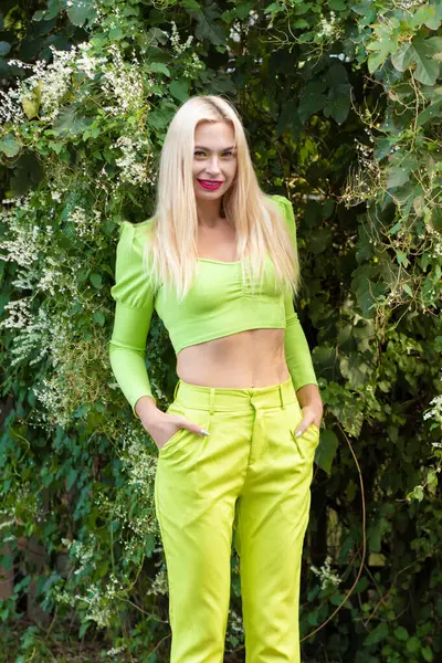 Sarı-yeşil takım elbiseli, uzun saçlı güzel bir sarışının portresi çiçekli yeşil çalıların arka planında.