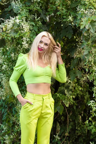 Sarı-yeşil takım elbiseli, uzun saçlı güzel bir sarışının portresi çiçekli yeşil çalıların arka planında.