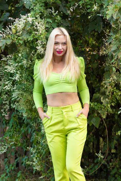 Sarı-yeşil takım elbiseli, uzun saçlı güzel bir sarışının portresi çiçekli yeşil çalıların arka planında.