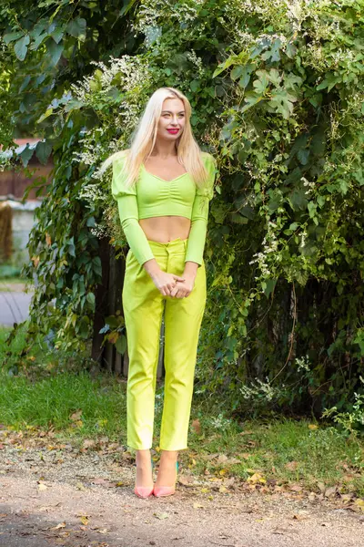 Sarı-yeşil takım elbiseli, uzun saçlı, güzel bir sarışının uzun boylu portresi ve çiçekli yeşil çalıların arka planında parlak yüksek topuklu ayakkabılar.