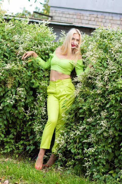 全长肖像一个漂亮的年轻金发姑娘穿着黄绿色的紧身衣和明亮的高跟鞋 背靠着开着花的绿色灌木丛 — 图库照片