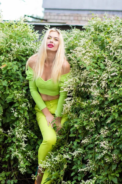 Sarı-yeşil takım elbiseli, uzun saçlı, güzel bir sarışının uzun boylu portresi ve çiçekli yeşil çalıların arka planında parlak yüksek topuklu ayakkabılar.